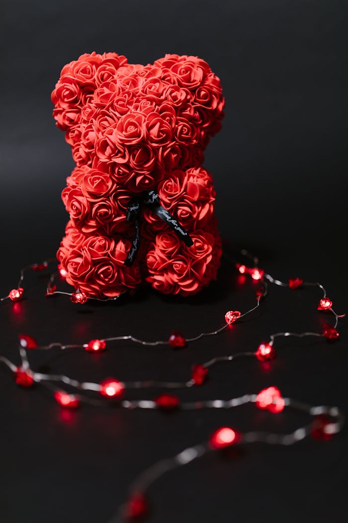 A decorative teddy bear made of red roses, surrounded by red string lights on a black background.